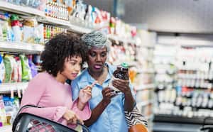 Dos mujeres afroamericanas comprando juntas en un supermercado. La más joven, de unos 20 años, lleva una cesta de la compra. La mayor, una mujer mayor de unos 60 años, sostiene una botella y están leyendo la etiqueta nutricional.