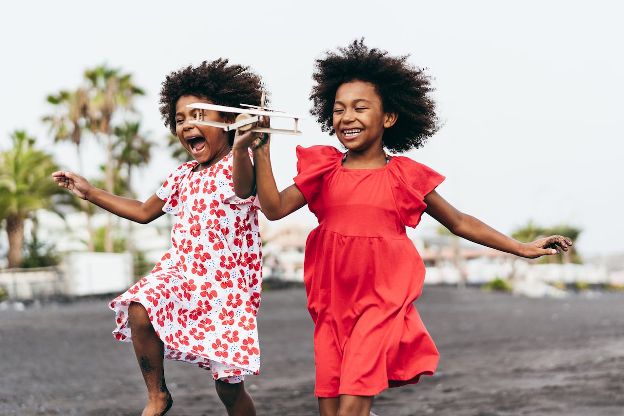 Hermanas gemelas afro corriendo en la playa mientras juegan con un avión de juguete de madera - Estilo de vida juvenil y concepto de viaje - Enfoque principal en la cara derecha del niño