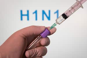 En esta ilustración fotográfica se ve una mano con un guante médico sosteniendo un tubo médico con una vacuna frente a una pantalla que dice "H1N1".